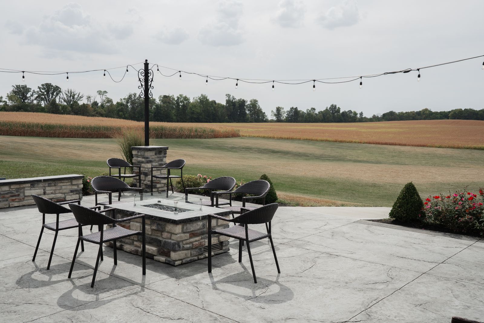 Stone fire-pit patio with string lighting overlooking open farmland at dusk